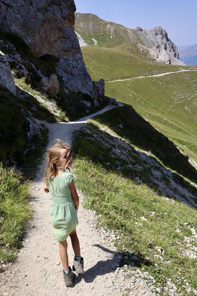 A young child in a light green dress and hiking boots walks along a winding dirt path on a sunny mountain slope